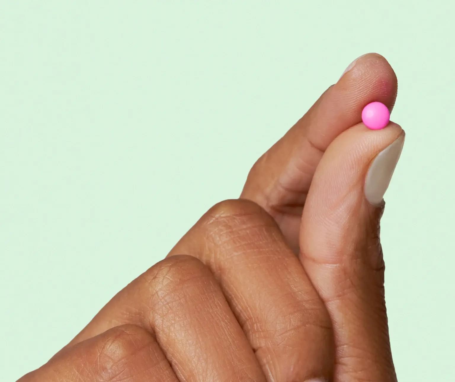 Fingers holding a small pink pill against a light green background.